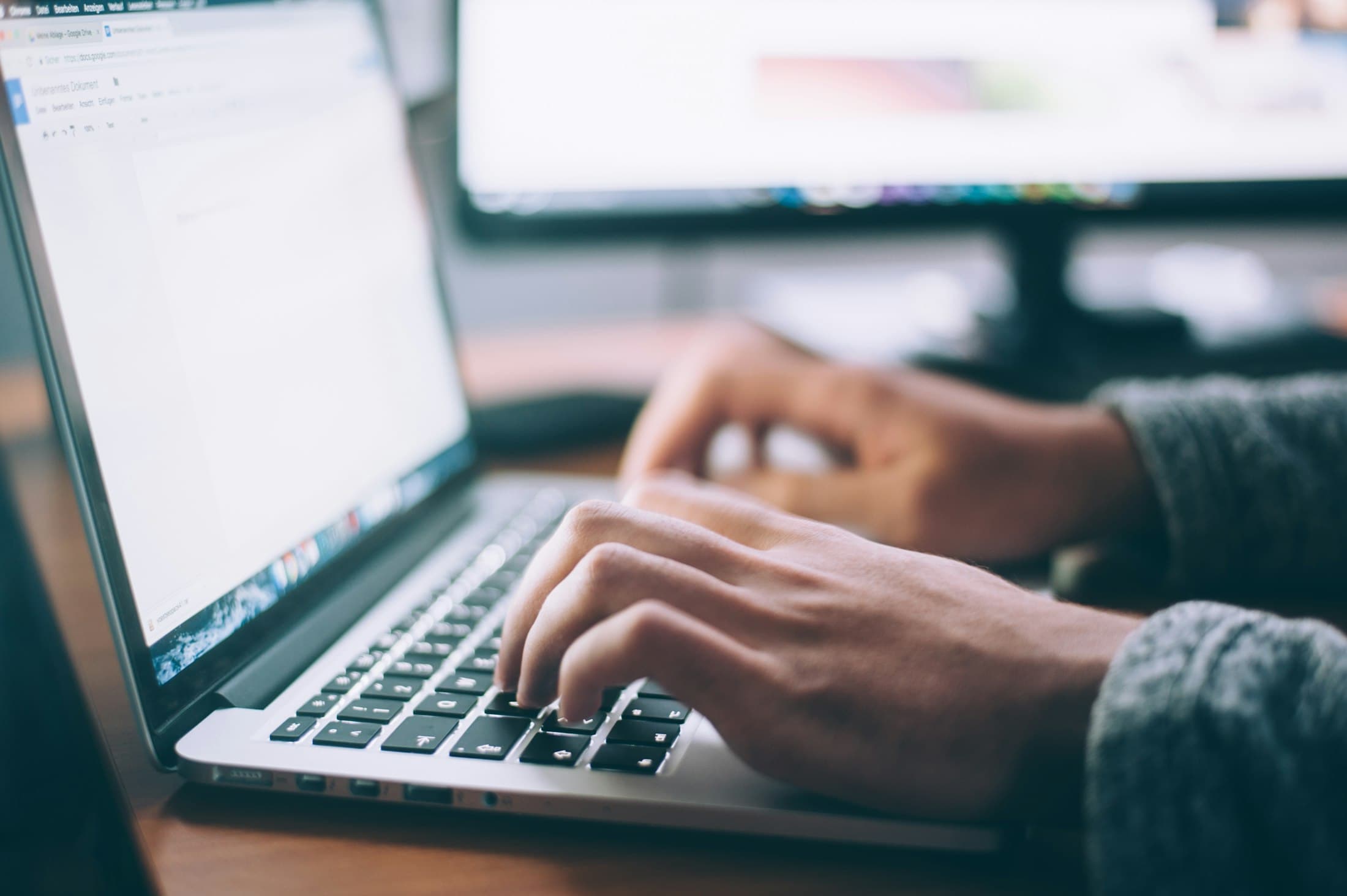 Hands typing on a laptop keyboard with a second monitor showing data in the background